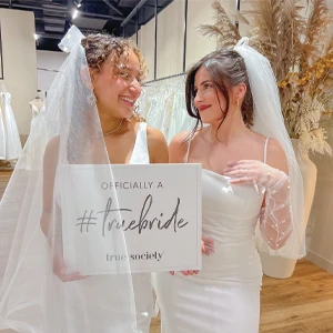 Two brides wearing white wedding gowns and veils stand together, smiling at each other. One bride holds a sign that reads 'Officially a #truebride true society.' They are in a bridal boutique with elegant decor and a rack of wedding dresses in the background