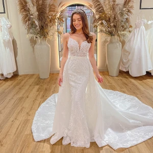 A bride wearing a lace wedding gown with a plunging neckline and intricate detailing stands in a bridal boutique. She smiles with her eyes closed and wears a tiara. The gown has a long, flowing train. The boutique features elegant decor, including large vases with pampas grass and other dried floral arrangements, and several wedding dresses hanging in the background