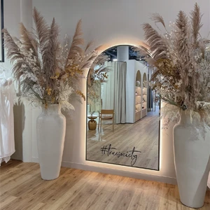 A bridal boutique features a large mirror with the hashtag '#truesociety' written on it. The mirror is framed by two large vases filled with pampas grass and other dried floral arrangements. The boutique has a warm and elegant decor, with wooden floors and a modern design. A chair and a small table are visible in the reflection