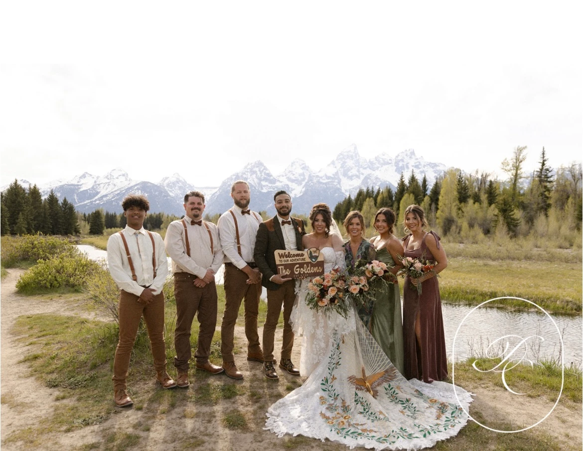 A bohemian bridal party poses against a backdrop of towering mountains, with the bride wearing a stunning gown adorned with nature-inspired embroidery.