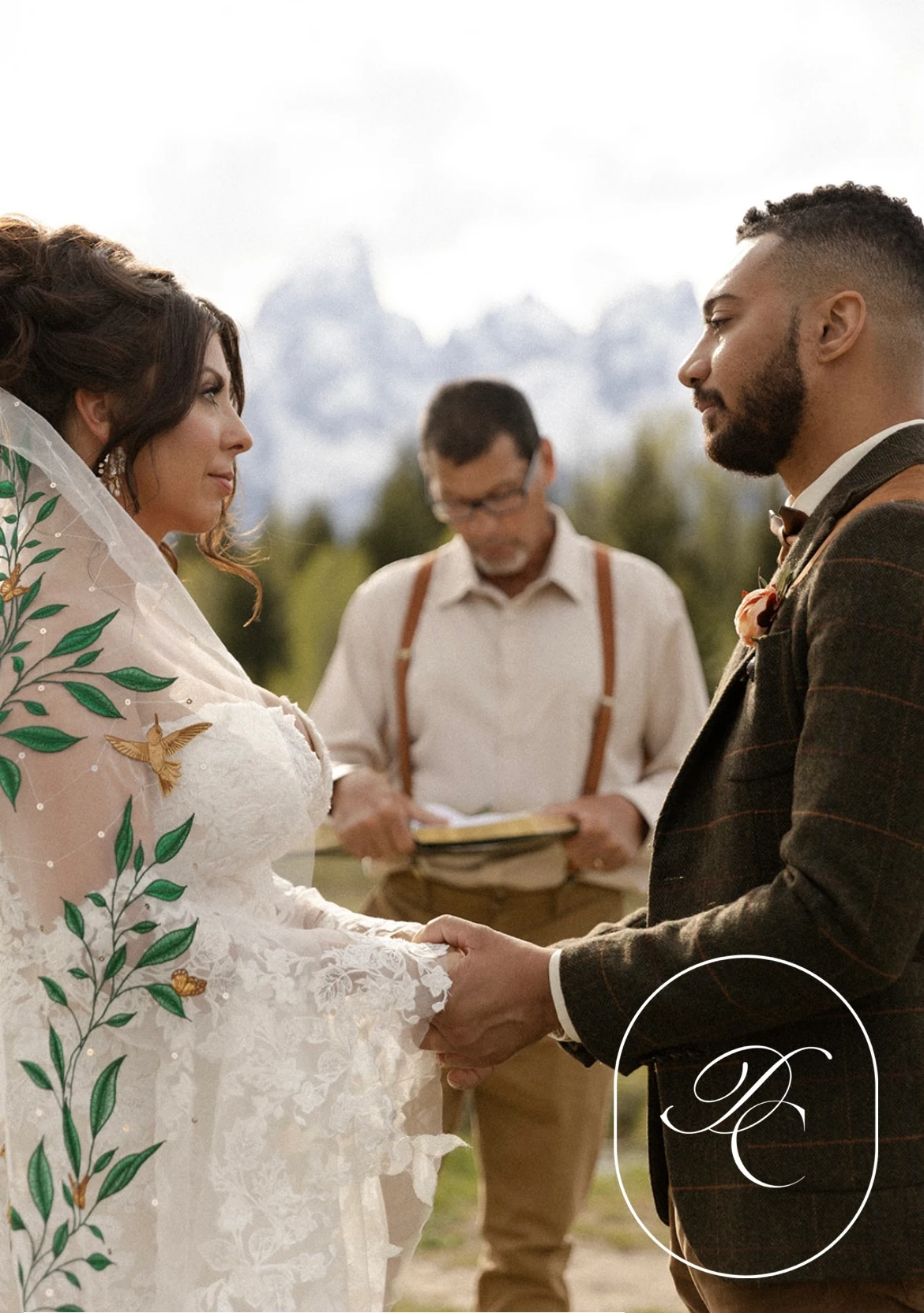 A outdoor ceremony with the bride wearing a veil beautifully adorned with leaf embroidery, and the groom in a rustic tweed suit, set against a mountain backdrop."