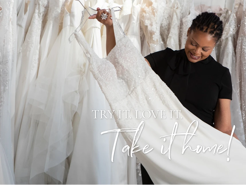 Smiling woman holding a white wedding dress in a bridal shop, surrounded by hanging wedding gowns. Text overlay reads 'Try it. Love it. Take it home!