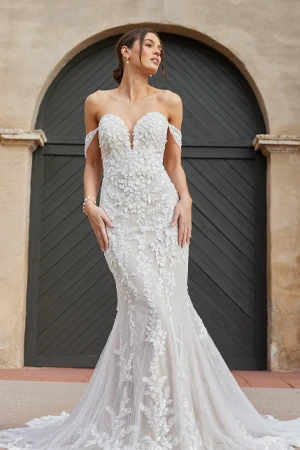 A bride poses elegantly in an off-the-shoulder mermaid-style wedding gown adorned with intricate lace and floral appliqués. The dress features a sweetheart neckline and a fitted silhouette that flares into a dramatic train. She wears a delicate bracelet and has her hair styled in a loose updo with soft tendrils framing her face. The background features an arched doorway with dark wooden doors, adding a timeless and romantic ambiance to the scene.