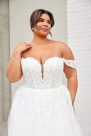 A bride poses confidently in an off-the-shoulder white wedding gown with a sweetheart neckline and intricate lace detailing. The dress features delicate floral appliqués and a flowing skirt. She has a radiant smile and her hair is styled in a soft updo with loose strands framing her face. The background features a modern white brick wall, adding a contemporary touch to the elegant scene.