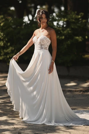 A bride poses outdoors in a strapless wedding gown with a sweetheart neckline and a flowing chiffon skirt. The bodice is adorned with delicate lace detailing, creating a romantic and elegant look. She holds the edge of her skirt, showcasing its soft movement. Her dark hair is styled in a loose updo, and she wears subtle jewelry. The background features lush greenery and dappled sunlight, adding to the serene and timeless feel of the scene.