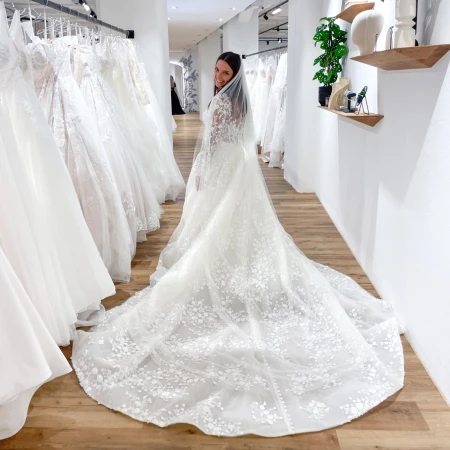 Braut in einem langärmeligen Spitzenbrautkleid mit einer kunstvollen floralen Schleppe, die in einer Brautboutique steht und über ihre Schulter lächelt. Sie ist von Reihen hängender Brautkleider umgeben, und ihr Schleier verleiht ihrem Look den letzten Schliff.