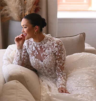 bride sitting on a couch wearing a high neck lace ballgown, 1517 by Martina Liana. She is looking out a window with her hand to her mouth smiling.