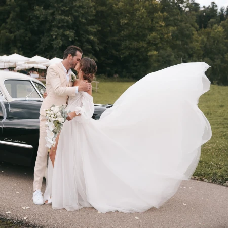 Braut und Bräutigam teilen einen intimen Moment im Freien neben einem Vintage-Auto. Das voluminöse Tüllkleid der Braut weht dramatisch im Wind, während sie sich umarmen. Der Bräutigam trägt einen hellen Anzug. Die Szene spielt vor einer Kulisse aus Grünflächen und einer parkähnlichen Umgebung.