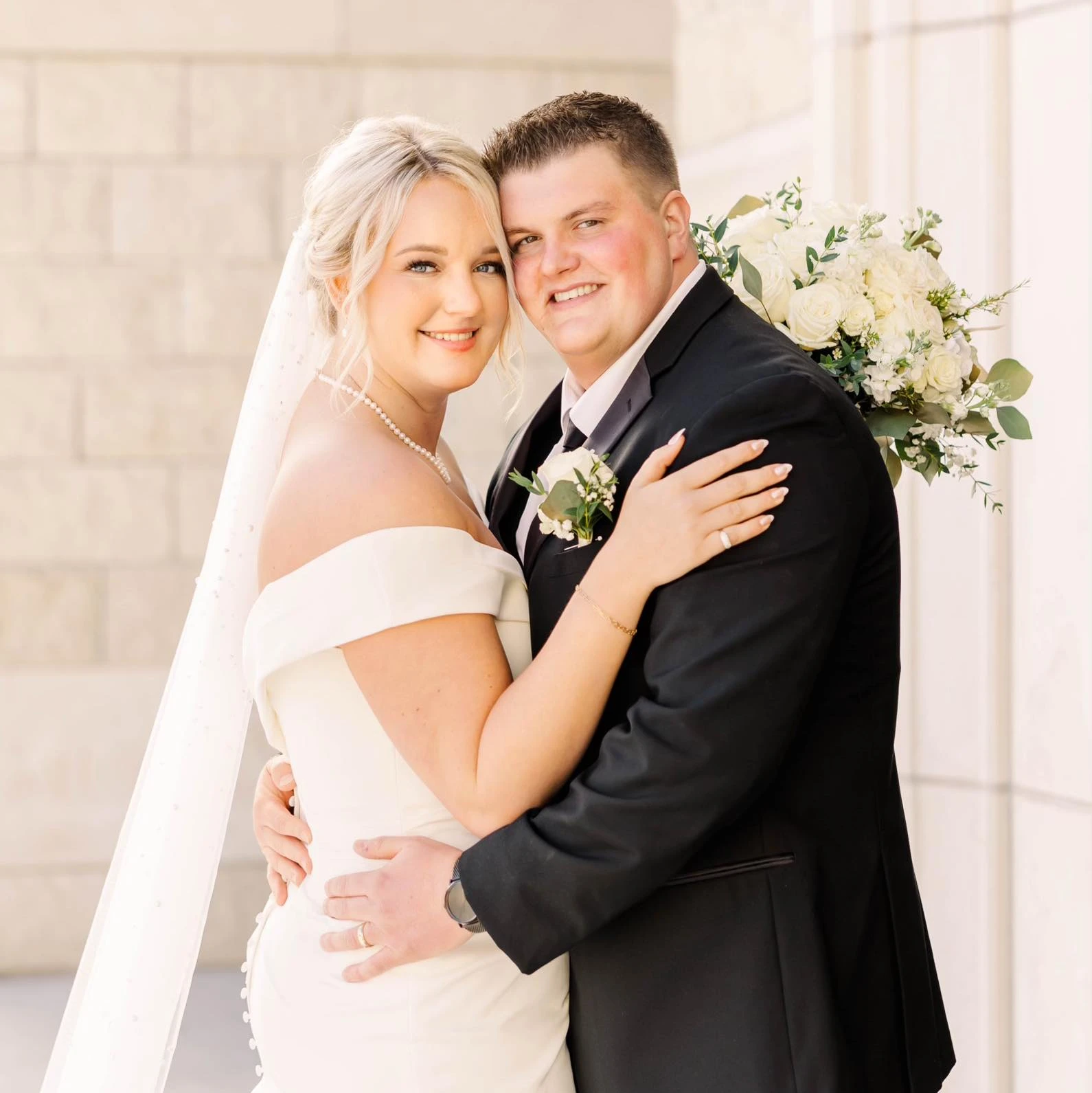 Groom and Bride embracing smiling. Bride is wearing an off the shoulder wedding gown with a veil. Groom is wearing a black tux.