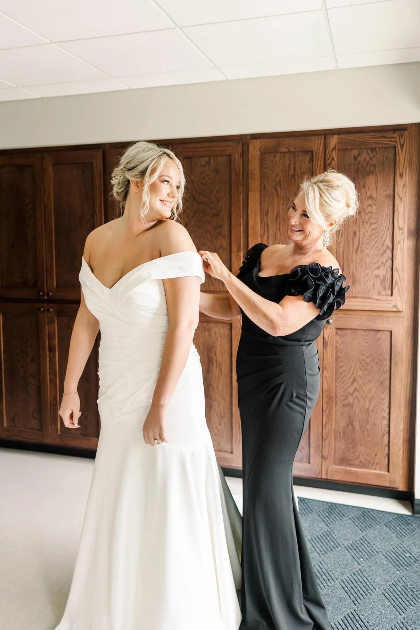 Mother of the bride wearing a off-the-shoulder black gown helping the bride zip up the back of her dress.