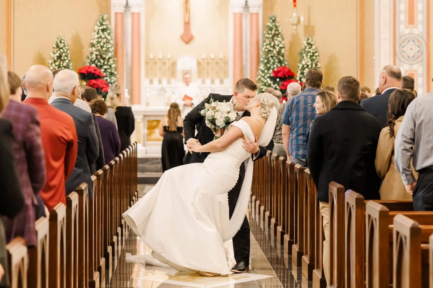 Groom kissing bride in the aisle of a church surrounded by wedding guests. 