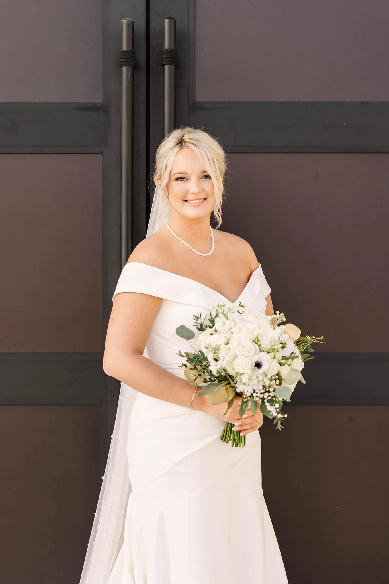 Bride standing wearing a off-the-shoulder wedding gown holding a bouquet of flowers.