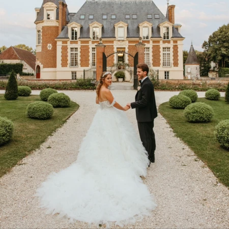 Braut und Bräutigam halten sich an den Händen und lächeln sich an, während sie auf einem Kiesweg vor einem prächtigen Schloss stehen. Die Braut trägt ein voluminöses, gerüschtes Brautkleid mit langer Schleppe, und der Bräutigam ist in einem schwarzen Anzug gekleidet. Gepflegte Gärten umgeben den Weg und verstärken die elegante, märchenhafte Atmosphäre.