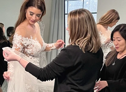 Bride in a lace wedding gown being assisted by two bridal stylists during a fitting session. The stylists are adjusting the gown’s train while the bride looks down at the dress. The scene takes place in a well-lit bridal boutique with a large window in the background.