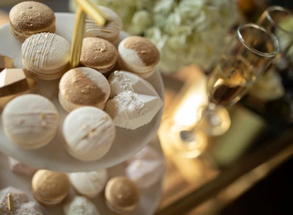 Elegant display of gold and white macarons and geometric-shaped desserts on a tiered serving tray, with glasses of champagne in the background. The setup creates a luxurious and celebratory atmosphere, perfect for a wedding or special event.