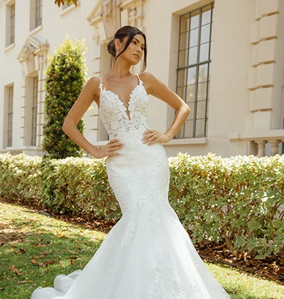 a bride is standing on a lawn wearing a lace mermaid wedding dress, D3484 by Essense of Australia. She has her hands on her hips and is glancing to the right.