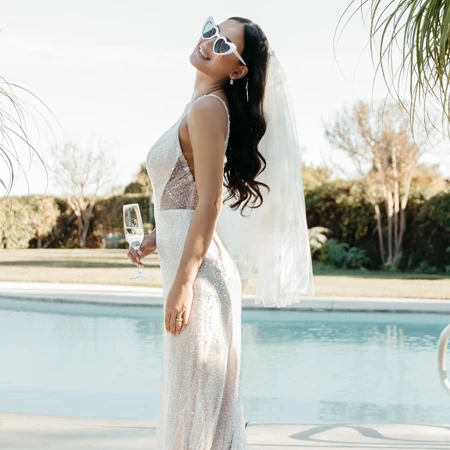 Braut in einem funkelnden Brautkleid und herzförmigen Sonnenbrillen, stehend am Pool mit einem Glas Champagner in der Hand. Sie lächelt und schaut nach oben, während ein langer Schleier hinter ihr weht und Palmen im Hintergrund sichtbar sind.