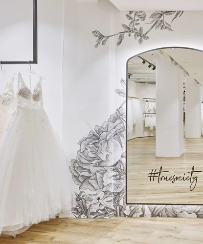 Brautboutique mit einem großen Spiegel, der von eleganten floralen Wandillustrationen umrahmt ist. Der Spiegel reflektiert das Innere der Boutique mit Reihen von Brautkleidern und hellem Holzboden. An der Wand ist der Hashtag '#truesociety' am unteren Rand des Spiegels angebracht, was eine moderne, social-media-freundliche Note hinzufügt.