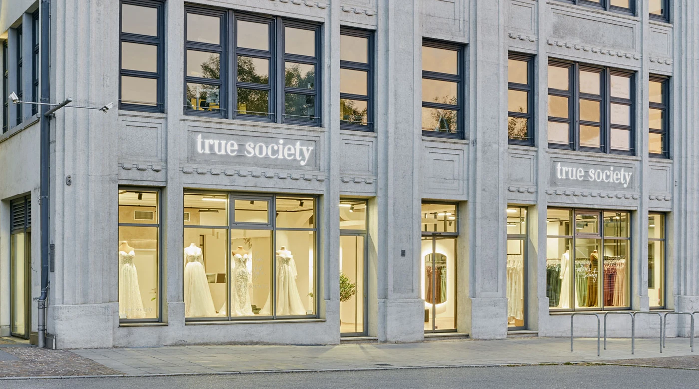 Außenansicht einer ‚True Society‘ Brautboutique mit großen Fenstern, die Brautkleider ausstellen. Das Gebäude hat ein modernes Design mit grauer Steinverkleidung und schwarz gerahmten Fenstern. Der Name der Boutique, ‚True Society‘, ist im oberen Teil des Gebäudes beleuchtet und schafft eine stilvolle und einladende Schaufensterfront.