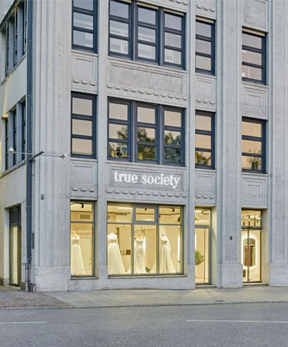 Außenansicht einer ‚True Society‘ Brautboutique mit großen Fenstern, die Brautkleider ausstellen. Das Gebäude hat ein modernes Design mit grauer Steinverkleidung und schwarz gerahmten Fenstern. Der Name der Boutique, ‚True Society‘, ist im oberen Teil des Gebäudes beleuchtet und schafft eine stilvolle und einladende Schaufensterfront.