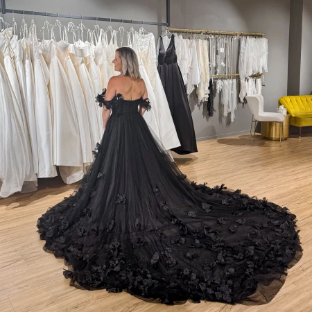 Bride wearing a dramatic black strapless wedding gown with a long, flowing train adorned with floral appliqués. She stands in a bridal boutique, facing away from the camera, with rows of white wedding dresses and a bright yellow chair in the background.