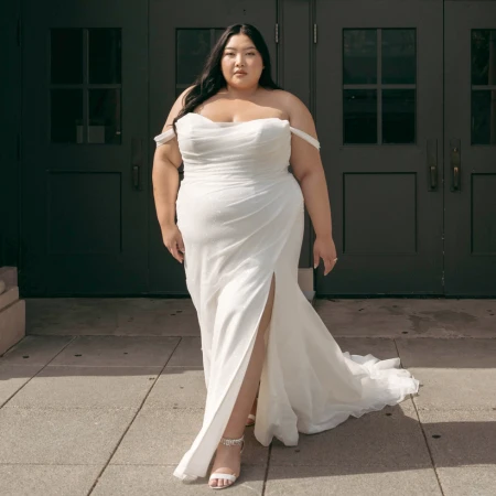 Bride wearing an off-the-shoulder, fitted white wedding gown with a thigh-high slit, confidently walking outdoors. The gown drapes elegantly behind her, and she pairs it with strappy heels. The background features dark double doors, adding contrast to the bride’s bright gown.