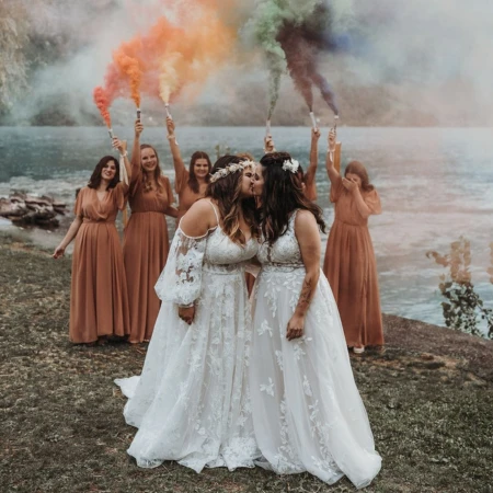 Two brides in lace wedding gowns share a kiss outdoors by a lake, surrounded by bridesmaids wearing earthy-toned dresses. The bridesmaids hold colorful smoke sticks in shades of orange, red, green, and blue, creating a vibrant and celebratory atmosphere against the natural landscape.