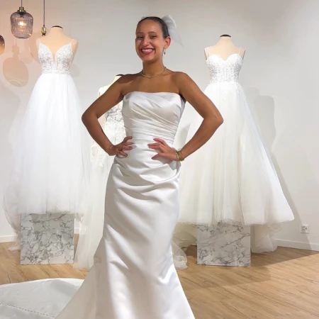 Bride in a sleek, strapless satin wedding gown, smiling and posing with her hands on her hips in a bridal boutique. Behind her are two mannequins dressed in lace and tulle wedding gowns displayed on marble stands, creating a chic and elegant setting.