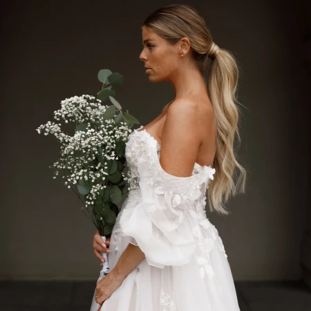 Bride in an off-the-shoulder wedding gown with delicate floral details, holding a bouquet of baby's breath and eucalyptus. She has her hair styled in a low, voluminous ponytail and gazes thoughtfully to the side against a dark, neutral background.