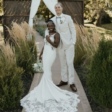 Bride and groom standing together outdoors in a garden setting with tall grasses and wooden doors draped in white fabric behind them. The bride wears a fitted lace gown with a long, detailed train and holds a small bouquet, while the groom wears a light suit with a bow tie and boutonniere, smiling with his arm around her.