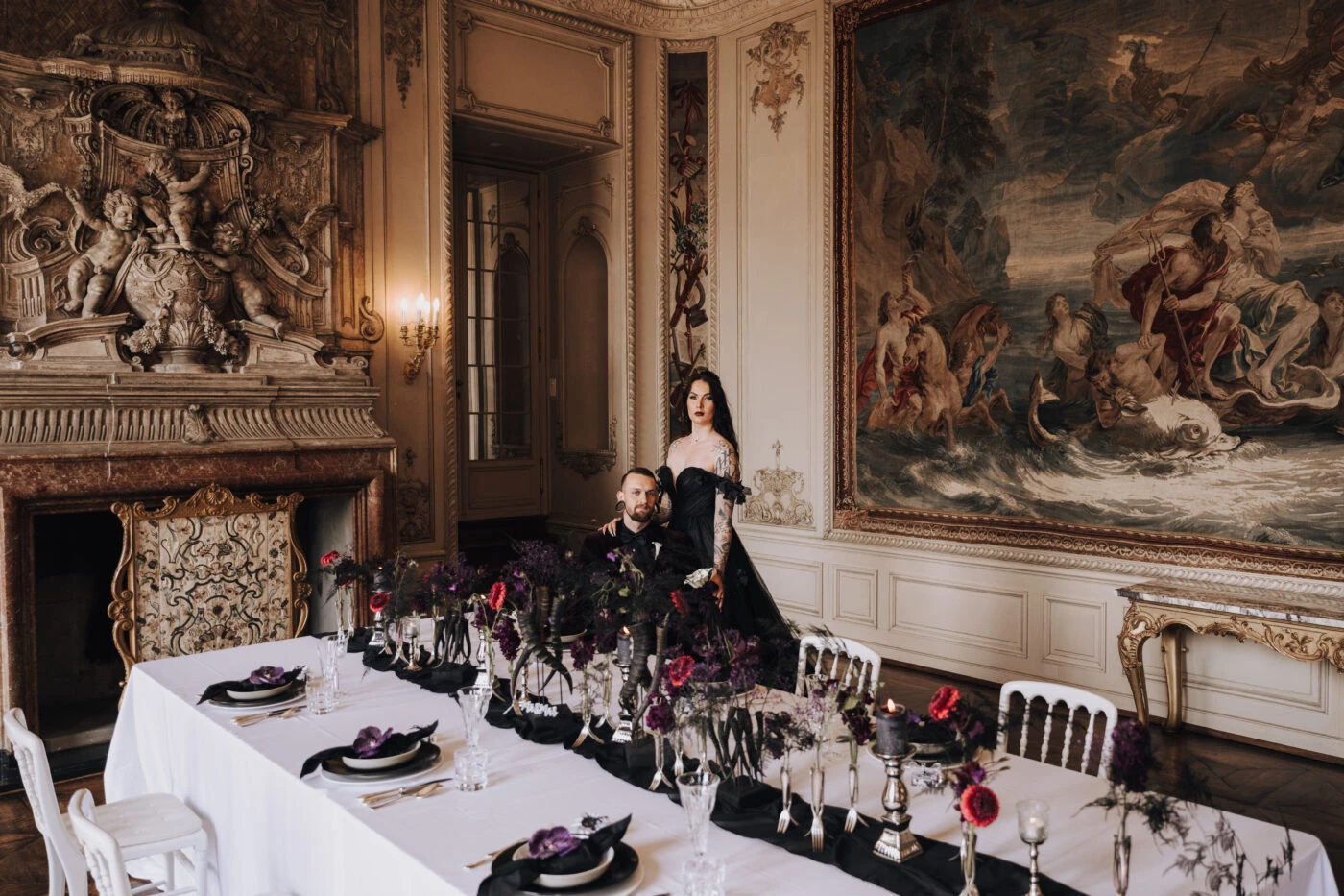 A couple in black wedding attire stands in an opulent room with ornate decor, a long table set with dark floral arrangements, and a large classical tapestry on the wall.