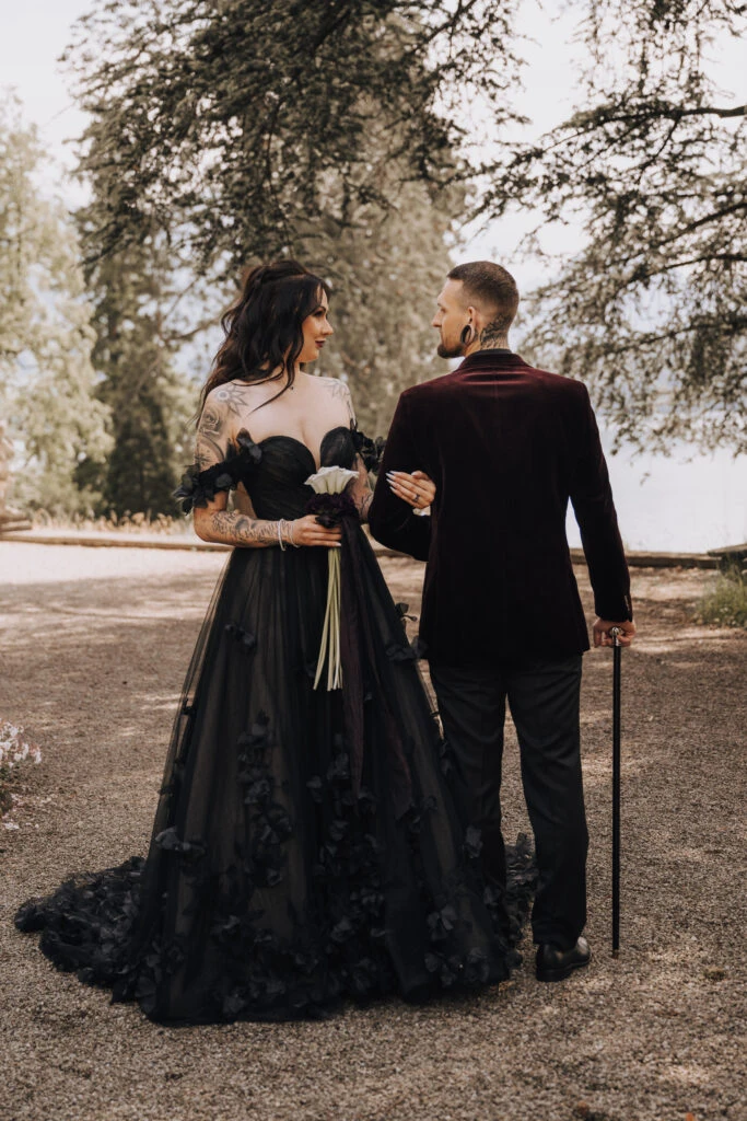 A couple in black wedding attire shares a quiet moment outdoors, with the bride holding a dark bouquet and the groom dressed in a velvet jacket and holding a cane.