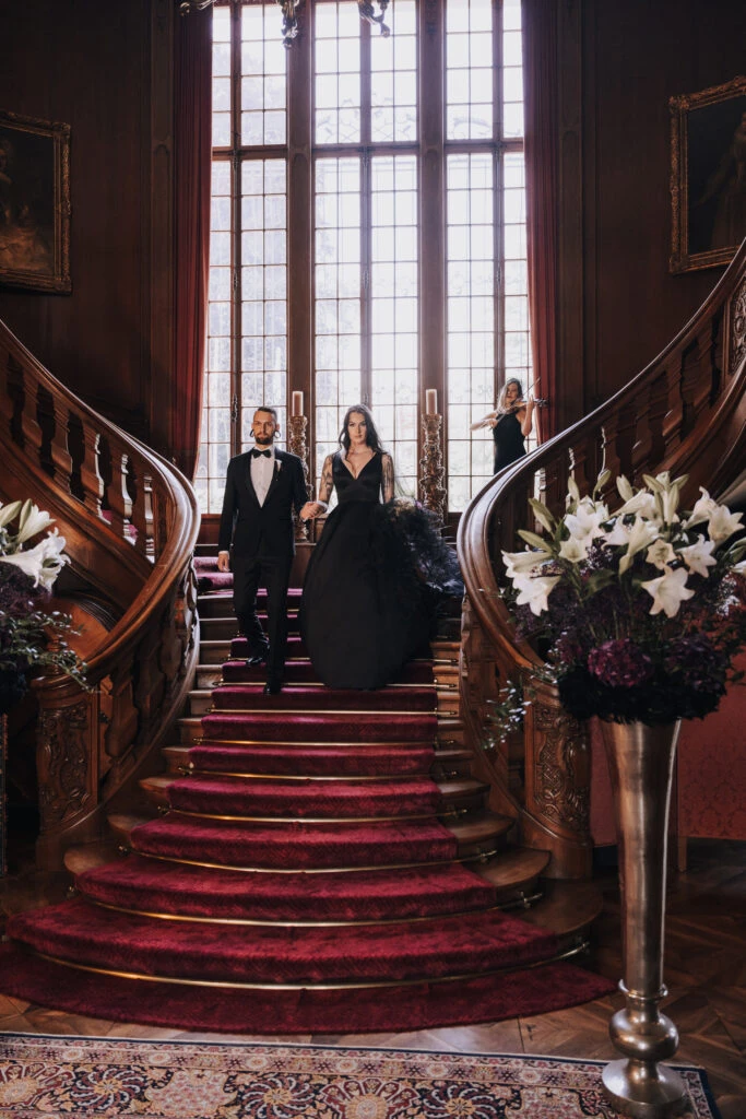 A couple descends a grand staircase, the bride in a black gown, the groom in a tux. Floral decor and a violinist in the background add to the romantic ambiance.