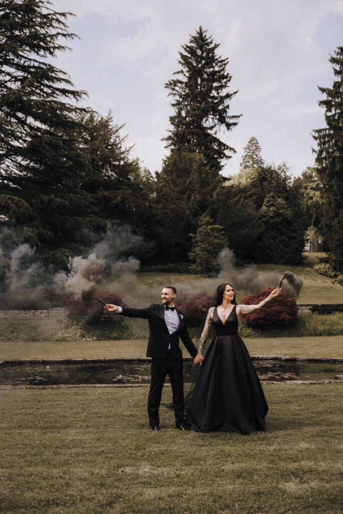 A couple in formal attire stands outdoors, holding smoke flares. The man wears a black tux, and the woman a black V-neck gown, with smoke swirling behind them.