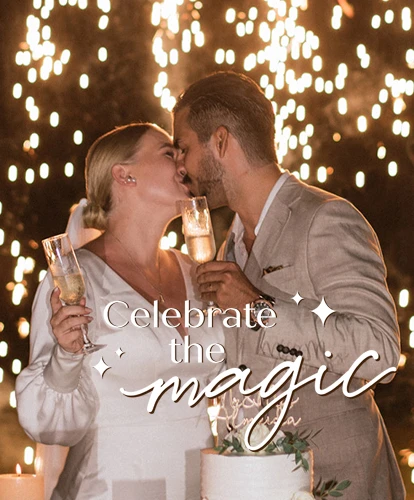 A newlywed couple shares a kiss while holding champagne glasses during a nighttime celebration with warm candlelight and sparkling lights in the background. A tiered white wedding cake adorned with flowers sits in the foreground, with the text 