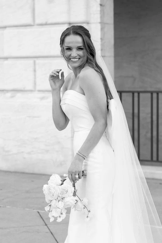 Bride laughing swearing a strapless wedding dress with a long tulle veil, holding a bouquet of flowers.