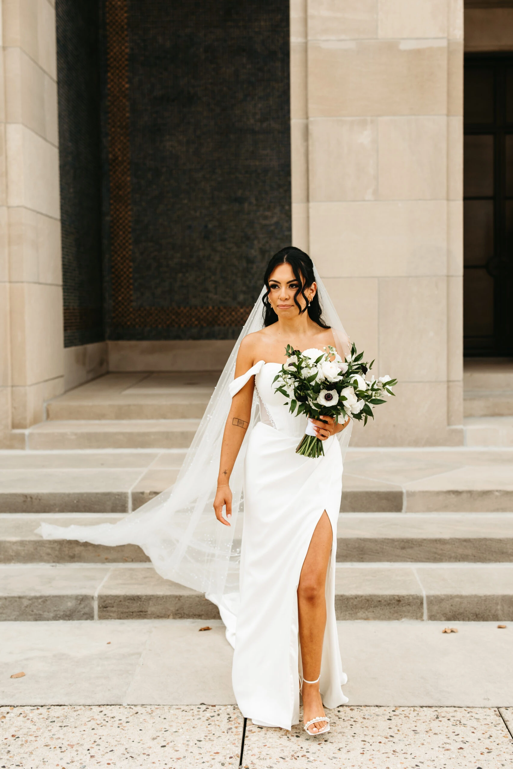 Bride walking down the strep outside a building wearing a off the should wedding dress and a cathedral veil. 