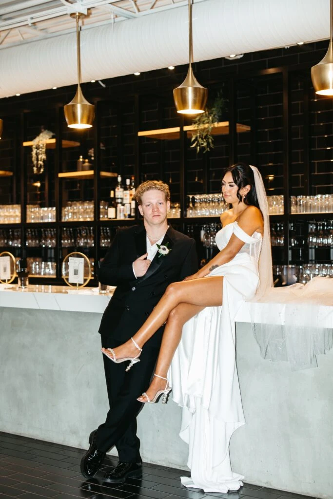 bride sitting on top of a bar table. While groom is leaning up against it. 