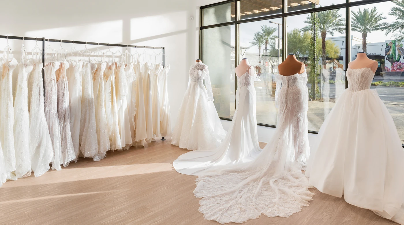 A bridal boutique display featuring a rack of intricately designed wedding dresses on hangers to the left, alongside mannequins showcasing elegant wedding gowns with long trains to the right. Sunlight streams through large windows, highlighting the delicate lace, tulle, and satin fabrics, while a view of palm trees and a street scene is visible outside. The space exudes sophistication and charm.