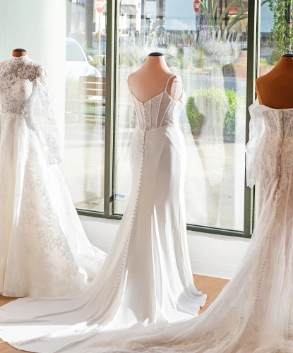 Close-up of mannequins in a bridal boutique showcasing three elegant wedding gowns with detailed lace and button-back designs. Each gown features long flowing trains and intricate detailing, illuminated by sunlight streaming through large windows. The outdoor view includes palm trees and a street scene, enhancing the boutique's bright and sophisticated atmosphere.