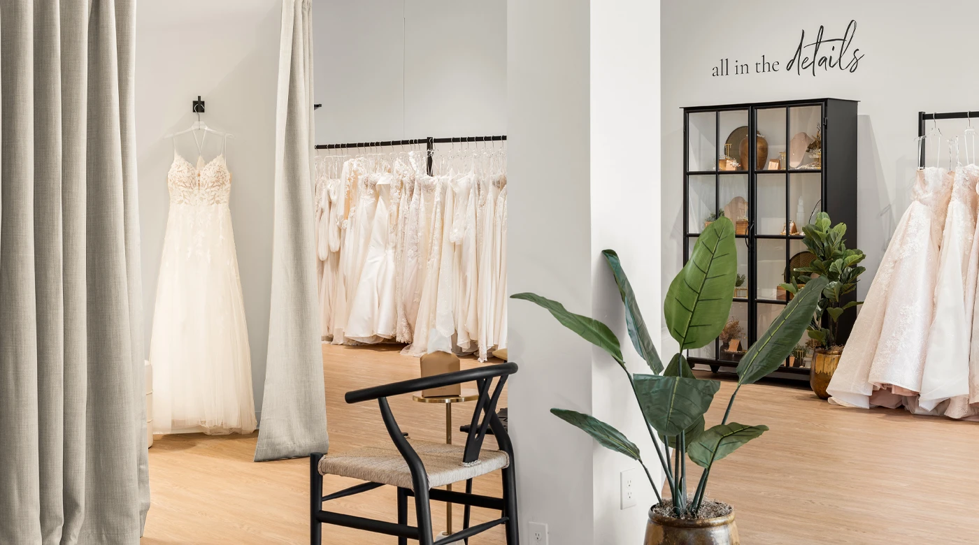 A cozy bridal boutique interior featuring a hanging lace wedding gown in a dressing area framed by neutral gray curtains. A black chair with woven seating and a large potted plant sit in the foreground, complementing the modern, serene decor. In the background, racks of wedding dresses and a black glass display cabinet labeled "all in the details" showcase decorative accents, creating an inviting and elegant shopping environment.