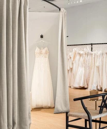 A charming bridal boutique showcasing a lace wedding gown hanging in a softly lit dressing area framed by gray curtains. A black chair with woven seating sits in the foreground, adding to the boutique's warm and elegant atmosphere. Behind the dressing area, racks of wedding dresses line the wall, creating an inviting space for brides-to-be.