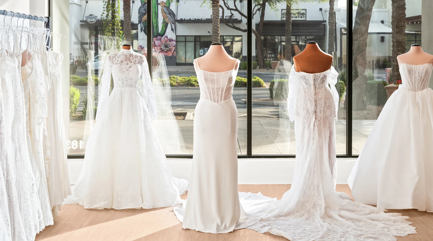 A bridal boutique display featuring mannequins dressed in elegant wedding gowns with intricate lace and satin details. The backdrop includes large floor-to-ceiling windows, allowing natural light to illuminate the dresses, and showcasing a view of palm trees and storefronts outside. The scene highlights the boutique's inviting and sophisticated atmosphere.