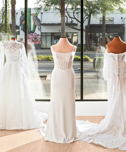 A close-up view of three mannequins in a bridal boutique, each adorned with elegant wedding gowns featuring lace and satin designs. The display is backlit by large windows, revealing a sunny exterior with trees and storefronts, adding a bright and inviting ambiance to the scene.