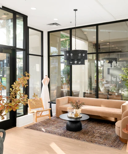 A cozy and elegant bridal boutique lounge area with a beige sofa and matching chair, centered around a black round coffee table adorned with decorative items. A lace bridal gown on a mannequin stands near large floor-to-ceiling windows that fill the space with natural light. The lounge is accented with a woven chair, a patterned rug, greenery, and a modern black chandelier, combining warmth and sophistication.