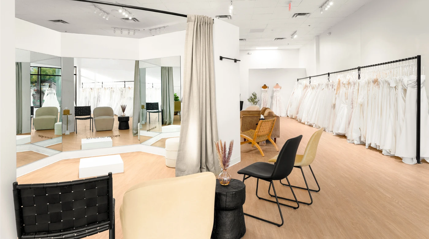 A modern bridal boutique interior showcasing a spacious fitting area with multiple mirrors and a white platform for dress fittings. Comfortable seating, including beige and black chairs, is arranged near a small black side table with decorative pampas grass. Rows of elegant white wedding dresses are displayed on racks along the right wall. The space features light wood flooring, neutral tones, and a welcoming, open layout.