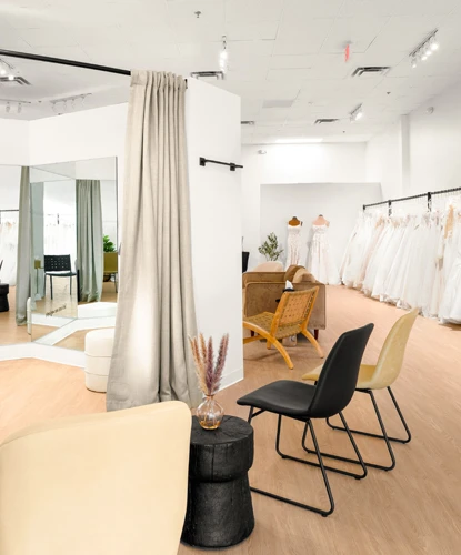 A bridal boutique featuring a fitting area with mirrors, a white fitting platform, and light beige curtains. The space includes modern seating with beige and black chairs near a small black side table holding decorative pampas grass. Rows of white wedding dresses are displayed on racks along the right side of the room. The interior has light wood flooring, high ceilings, and bright lighting for a welcoming atmosphere.