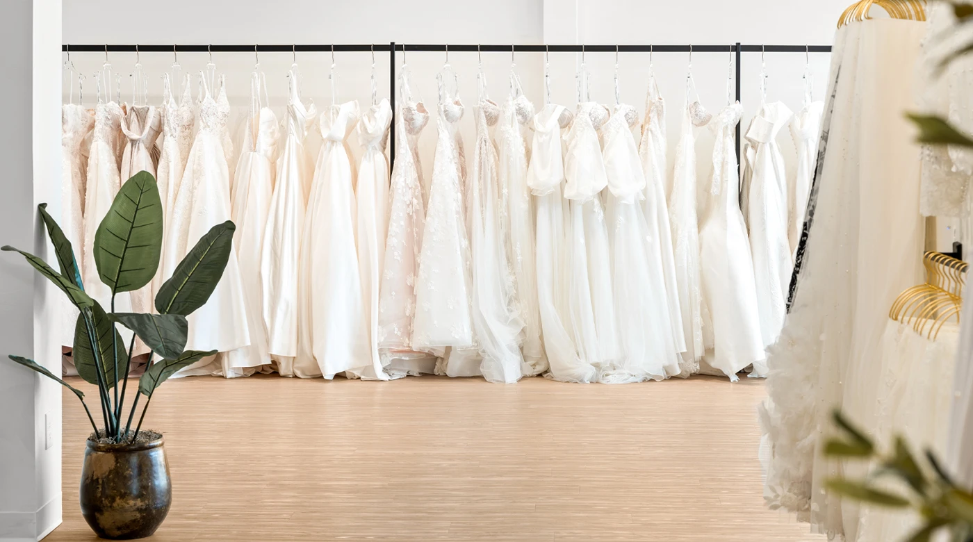 A display of elegant white and ivory wedding dresses hanging on racks in a bridal boutique. The gowns feature a variety of designs, including lace, satin, and tulle, some with intricate details like embellishments and bows. A large green potted plant adds a natural touch in the foreground, complementing the modern and minimalist interior with light wood flooring and soft lighting.