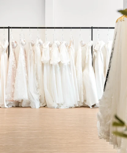 A selection of white and ivory wedding dresses hanging neatly on a rack in a bridal boutique. The dresses showcase various styles and textures, including lace, tulle, and satin, with intricate details such as bows and embellishments. Part of another dress rack is visible in the foreground on the right, creating depth within the modern, well-lit space with light wood flooring.