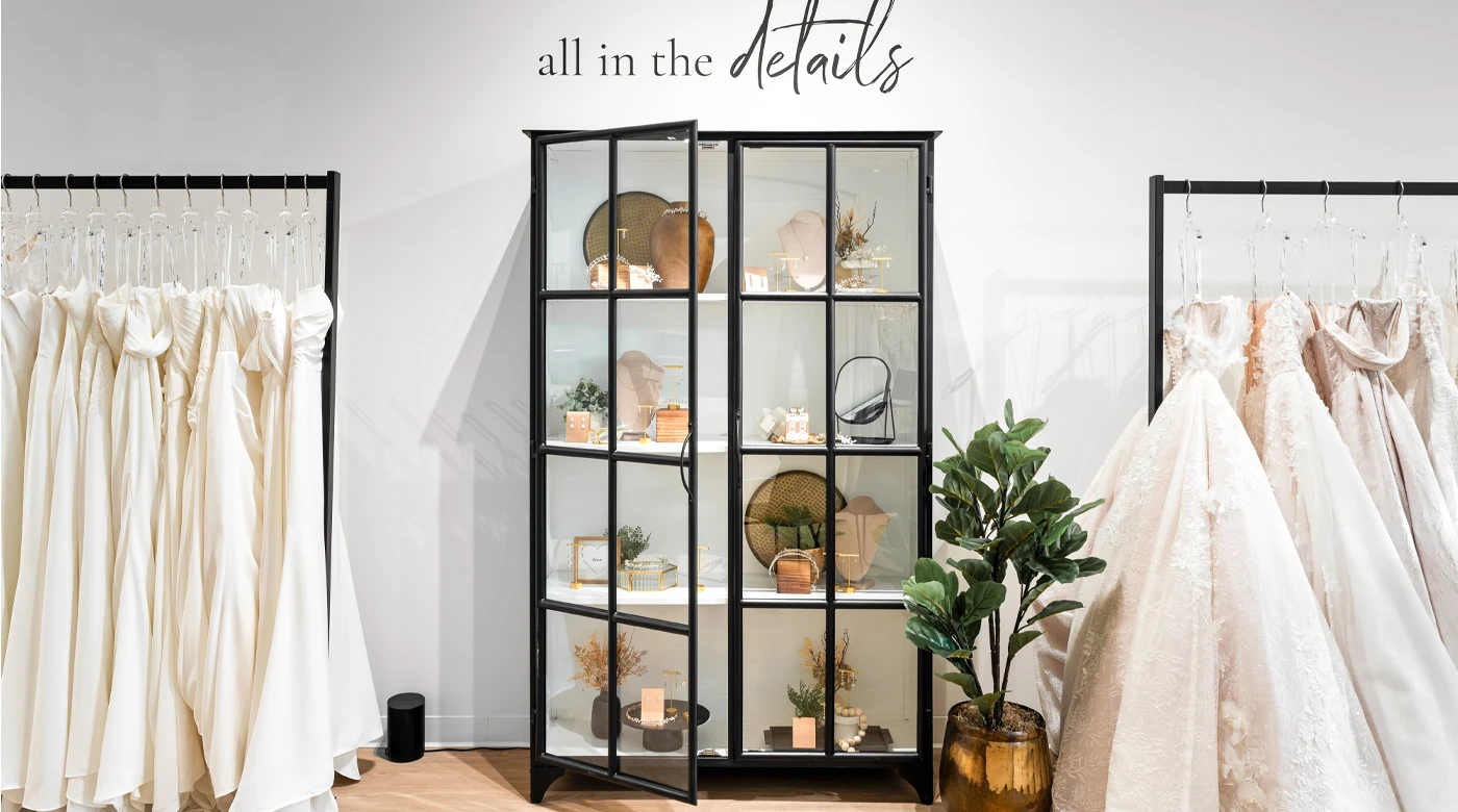 A modern bridal boutique featuring a sleek black display cabinet filled with curated wedding accessories, such as jewelry, hairpieces, and decor. On either side of the cabinet are racks of elegant wedding dresses in shades of white and blush, showcasing various textures and styles. A gold planter with lush green foliage adds a touch of natural decor to the scene, with the phrase 